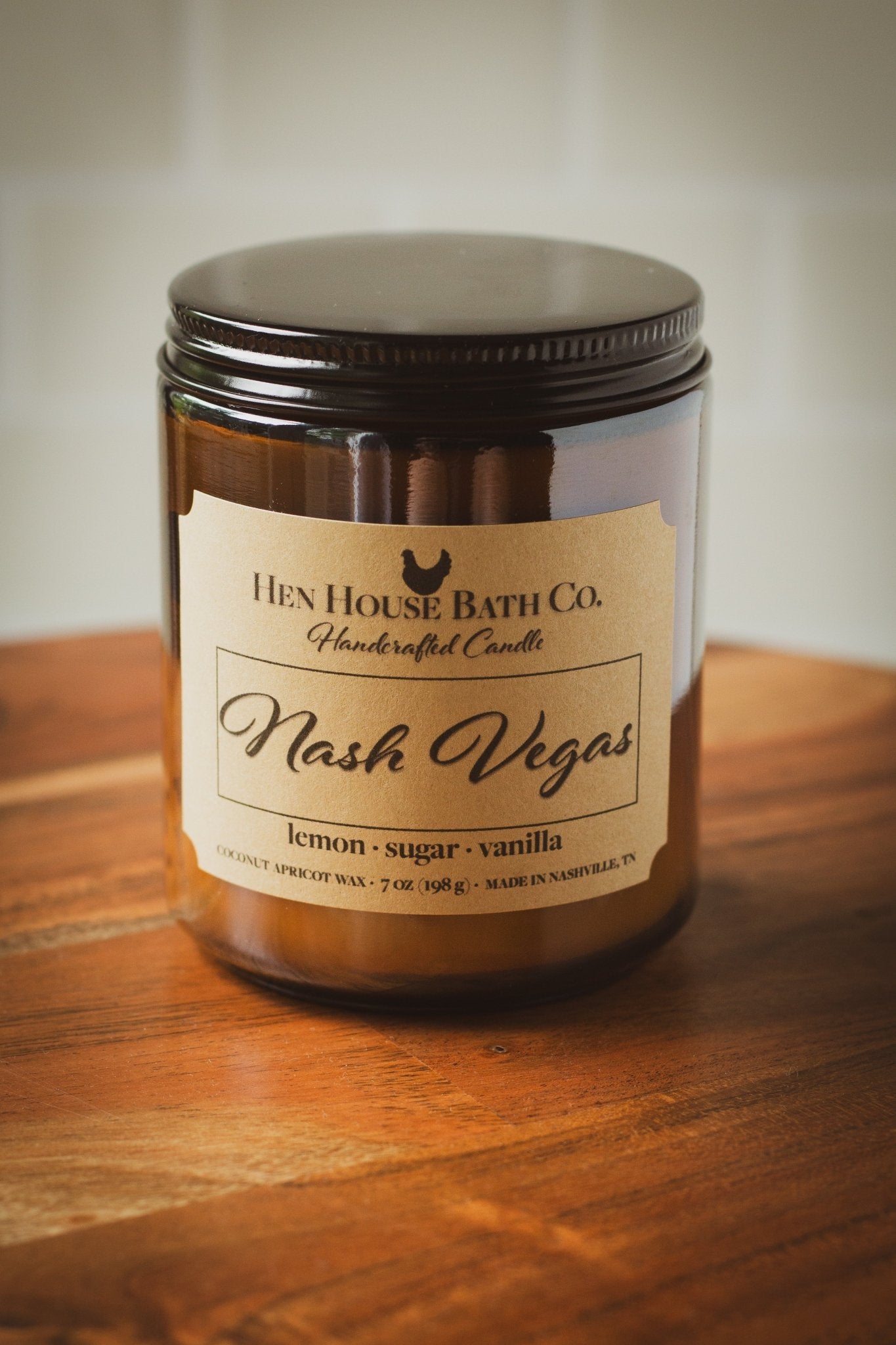 Amber glass candle jar with a kraft label reading “Hen House Bath Co. Handcrafted Candle — Nash Vegas.” Scent notes listed below as “lemon · sugar · vanilla.” The candle sits on a warm wooden surface, photographed in soft natural light.