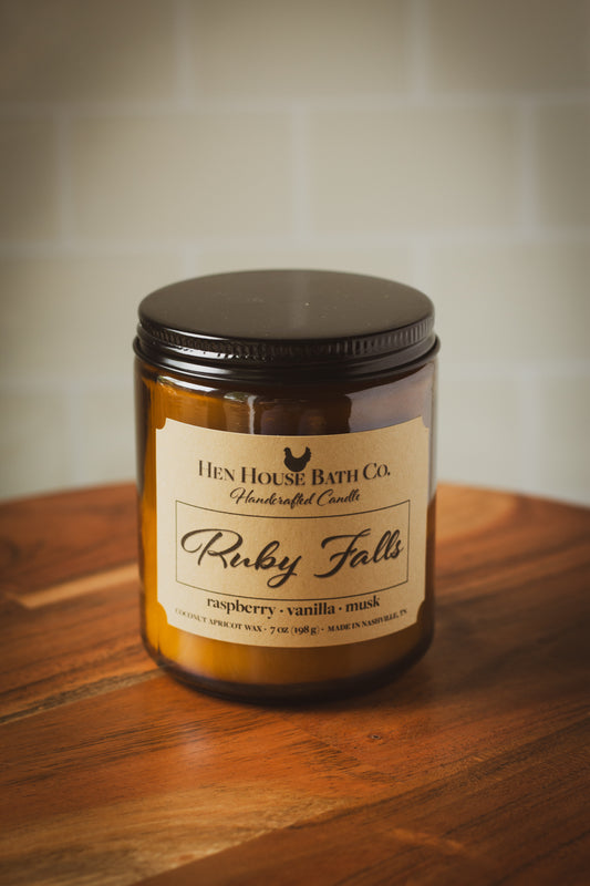 Amber glass candle jar with the lid closed. The label reads “Hen House Bath Co. Handcrafted Candle — Ruby Falls.” Scent notes listed as “raspberry · vanilla · musk.” The candle is placed on a wooden surface with a softly blurred, light-colored tile background.
