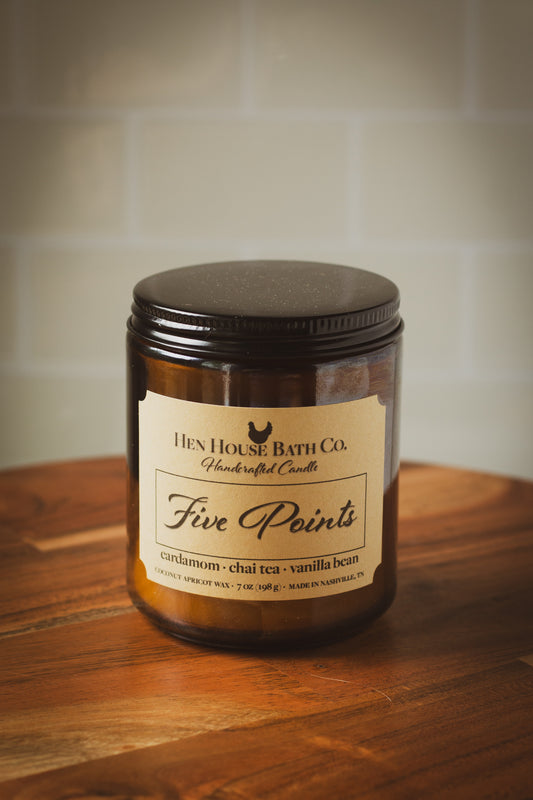 Amber glass candle jar with kraft label reading “Five Points” from Hen House Bath Co., sitting on a wooden surface with a neutral tile background.