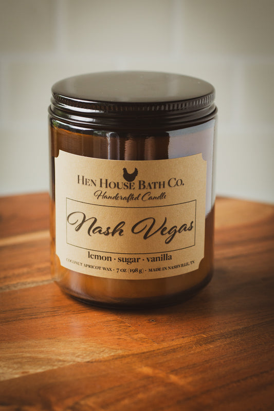 Amber glass candle jar with a kraft label reading “Hen House Bath Co. Handcrafted Candle — Nash Vegas.” Scent notes listed below as “lemon · sugar · vanilla.” The candle sits on a warm wooden surface, photographed in soft natural light.
