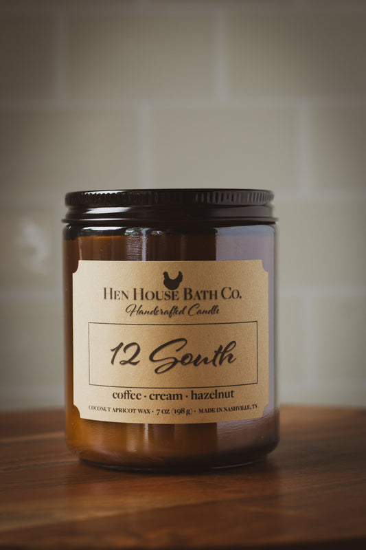 Amber glass candle jar with kraft label reading “12 South” from Hen House Bath Co., sitting on a wooden surface with a neutral tile background.