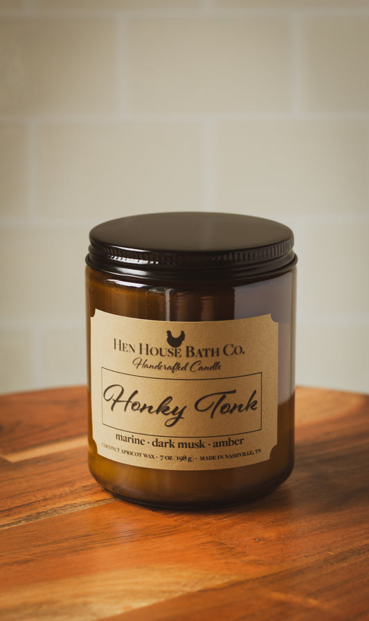 Amber glass candle jar with the lid closed. The label reads “Hen House Bath Co. Handcrafted Candle — Honky Tonk.” Scent notes listed as “marine · dark musk · amber.” The candle sits on a wooden surface with a softly blurred, light tile background.