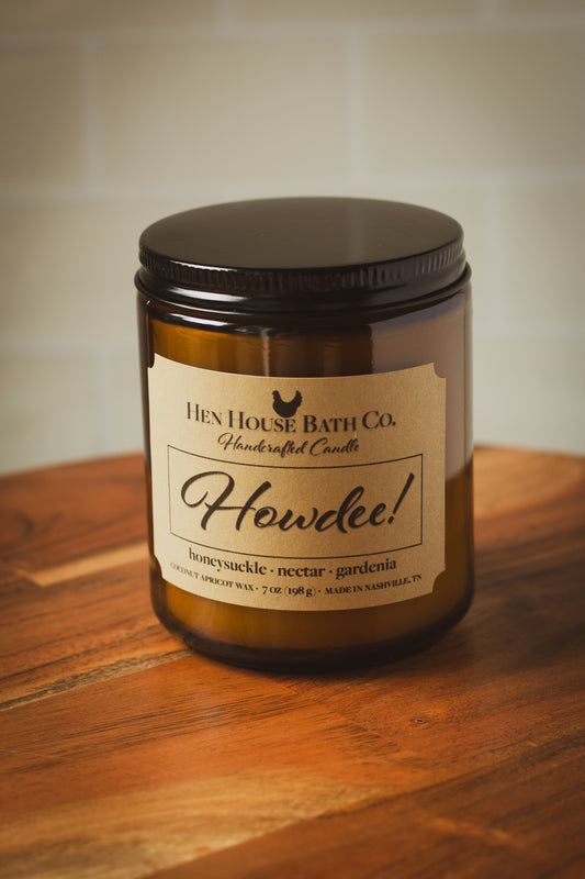 Amber glass jar candle with a kraft paper label reading “Hen House Bath Co. Handcrafted Candle — Howdee!” with scent notes of honeysuckle, nectar, and gardenia. The candle is sitting on a wooden surface with a light tiled background.
