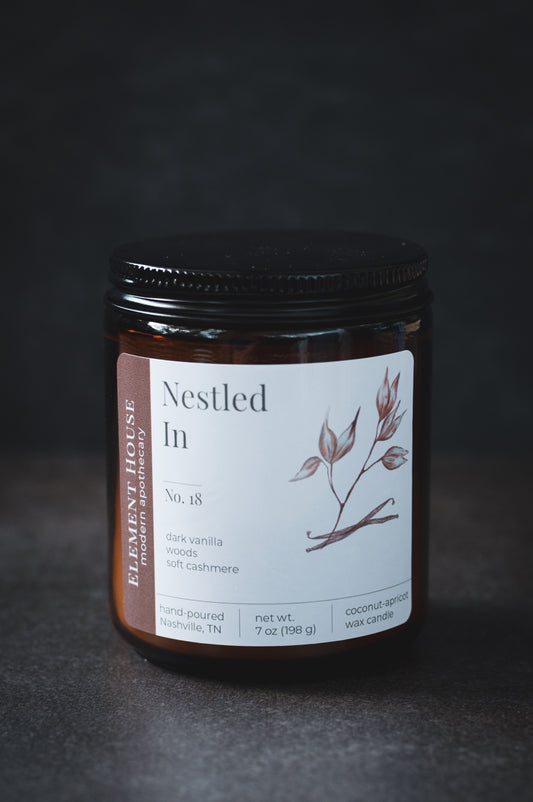 Nestled In candle in an amber glass jar with a black lid, featuring an Element House label with a botanical branch illustration, photographed against a dark textured background.