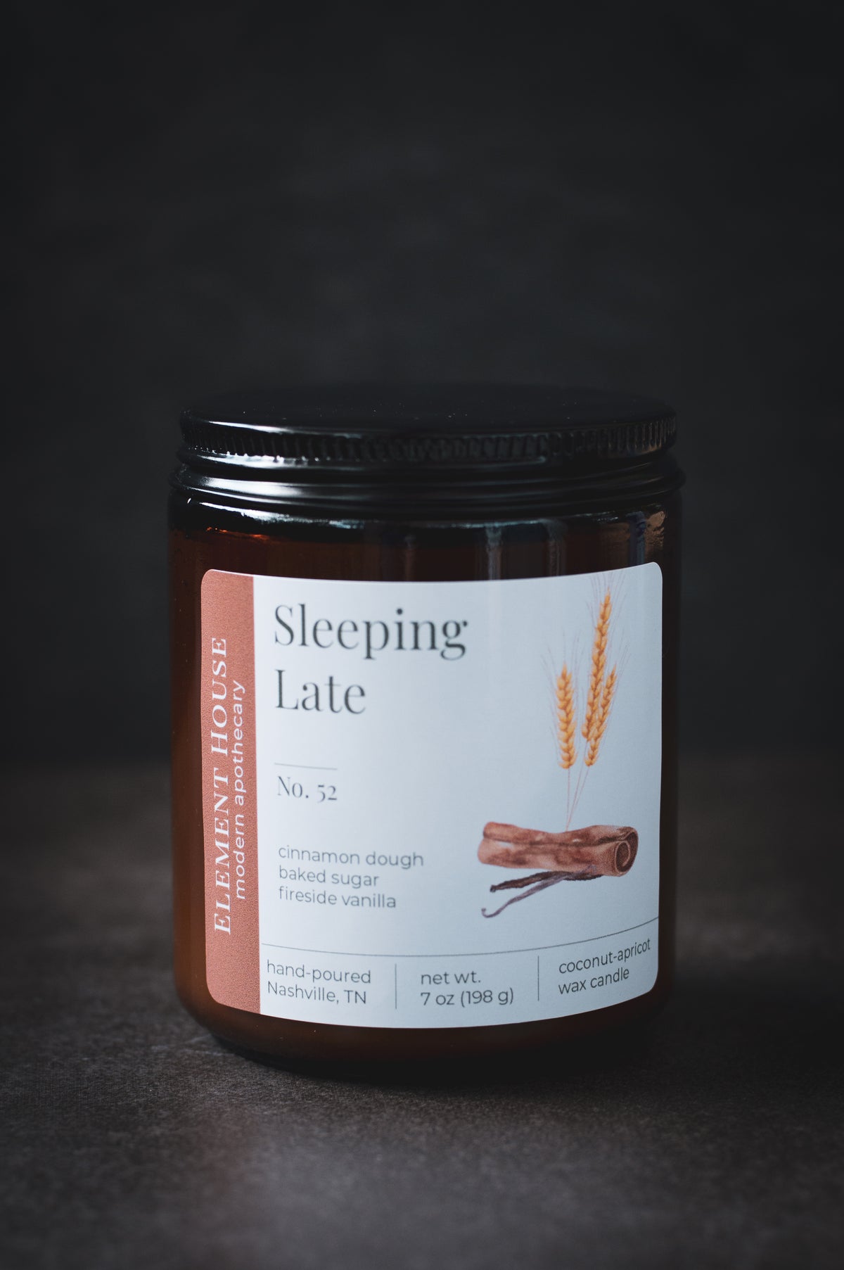 Sleeping Late candle in an amber glass jar with a black lid, featuring an Element House label with cinnamon sticks and wheat illustrations, photographed against a dark background.