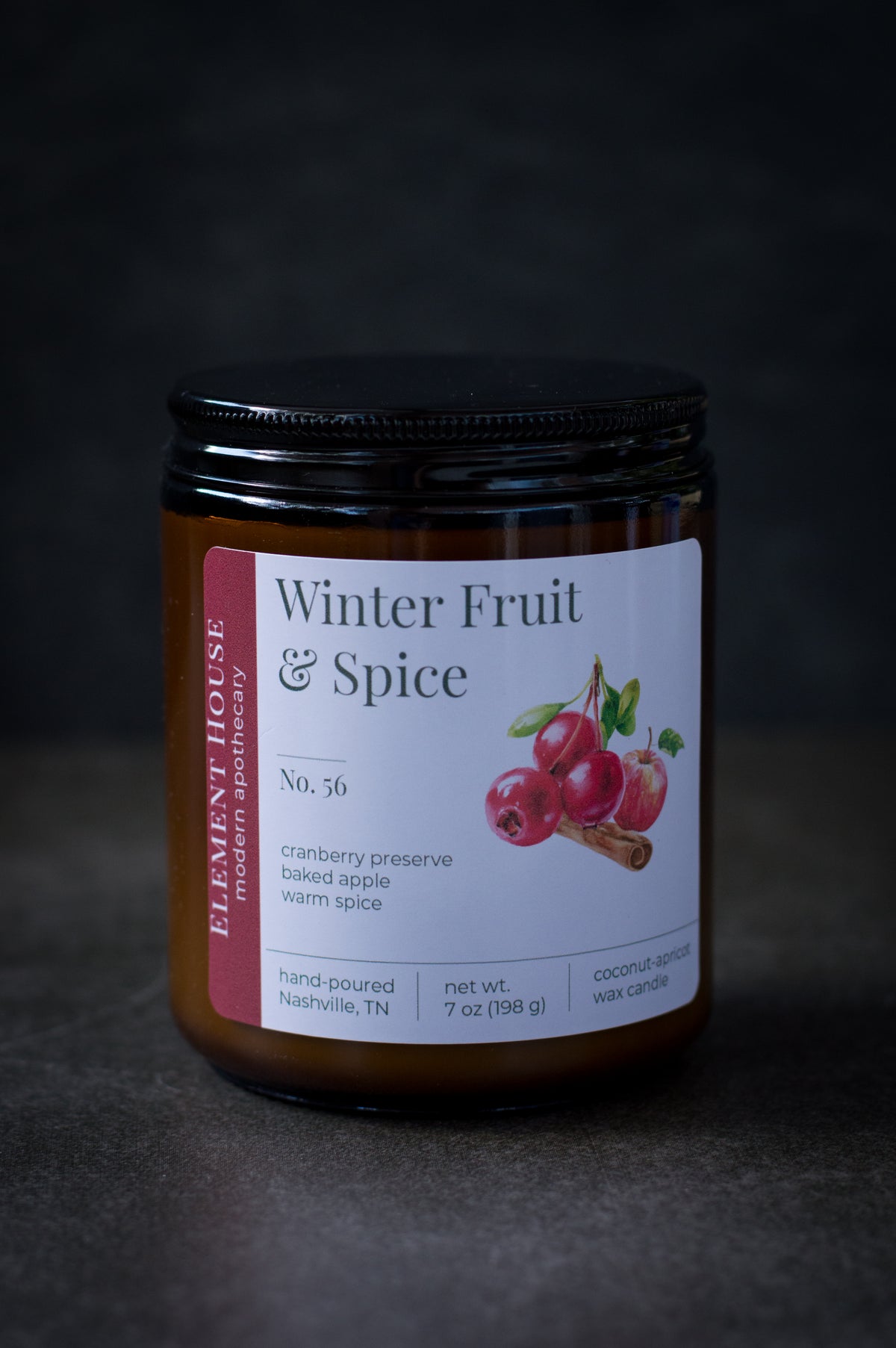 Winter Fruit & Spice candle in an amber glass jar with a black lid, featuring an Element House label with cranberry, apple, and cinnamon illustrations, photographed against a dark background.