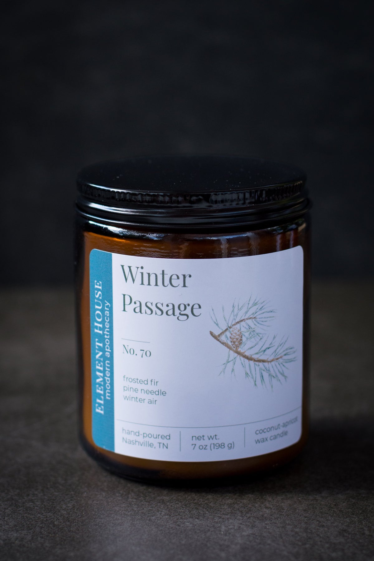 Winter Passage candle in an amber glass jar with a black lid, featuring an Element House label with an evergreen branch illustration, photographed against a dark textured background.