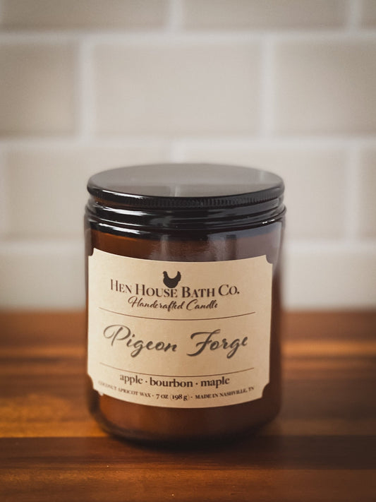 Hand-poured candle inspired by cozy cabin nights in Pigeon Forge with warm notes of apples, maple, and bourbon by Hen House Bath Co. in amber jar on rustic background.