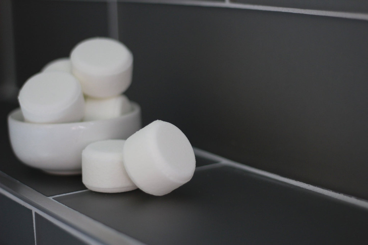 White round shower steamers stacked in a ceramic bowl on a dark bathroom tile ledge, minimalist and clean style.
