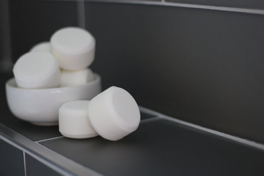 White round shower steamers stacked in a ceramic bowl on a dark bathroom tile ledge, minimalist and clean style.